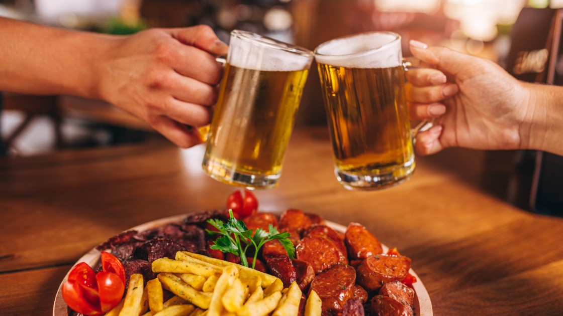 Two people clinking beer glasses over a platter of assorted food including sausages, fries, and tomatoes.